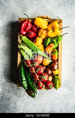 Ansicht von oben auf eine Holzkiste mit Kirschtomaten auf der Rebe, Okra, roten, grünen und gelben Chilischoten gefüllt Stockfoto