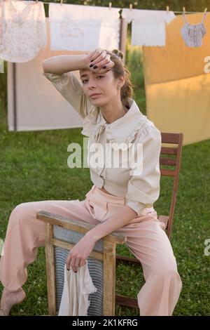 Wäschereitag. Eine Frau hängt Bettwäsche und Handtücher auf einem Baum im Hof eines Dorfhauses. Sommerhaus-Konzept. Stockfoto