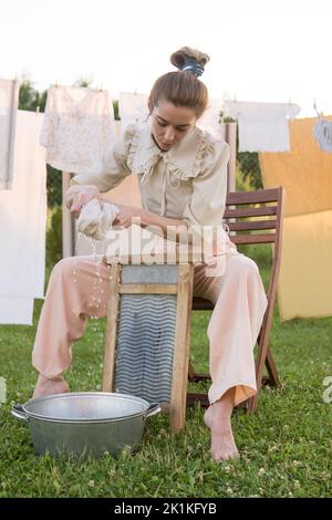 Wäschereitag. Eine Frau hängt Bettwäsche und Handtücher auf einem Baum im Hof eines Dorfhauses. Sommerhaus-Konzept. Stockfoto