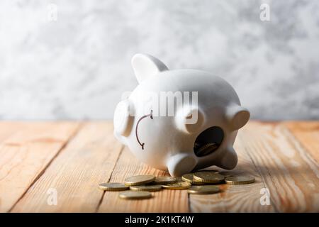 Ein trauriges Sparschwein hinter einem Stapel von Euro-Münzen, das den Fall der Geldwerte symbolisiert. Weiße Geldbox mit goldenen Münzen. Krisenkonzept Stockfoto