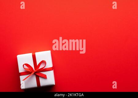 Kreatives Postkarten-Design zu Weihnachten und Valentinstag mit weißer Geschenkbox und roter Schleife auf rotem Papierhintergrund. Valentinstag und Liebeskonzept Stockfoto