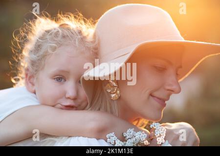 Seitenansicht der erstaunlichen Familie, die im Sommer im Wald steht. Junge lächelnde Mutter gibt Huckepack Fahrt zu kleinen Mädchen. Stockfoto