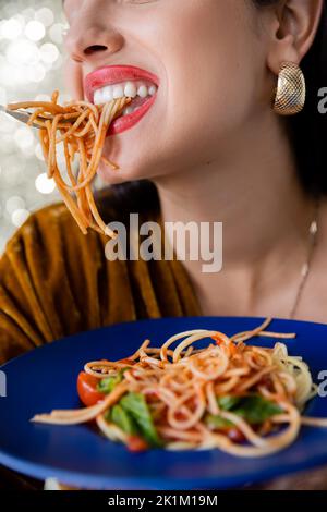 Teilansicht einer Frau mit roten Lippen und goldenem Ohrring beim Essen von Pasta aus blauem Teller auf glänzendem Hintergrund, Stockbild Stockfoto