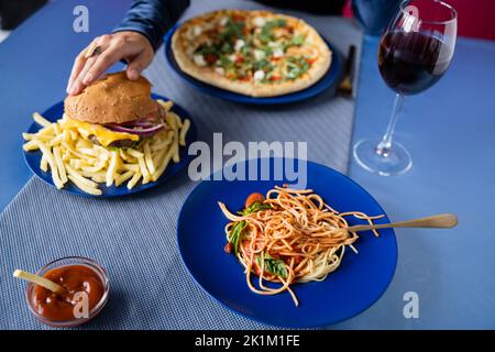 Teilansicht einer Frau, die Burger in der Nähe von Weinglas und Teller mit Pasta und Pizza auf blauem Tisch nahm, Stockbild Stockfoto