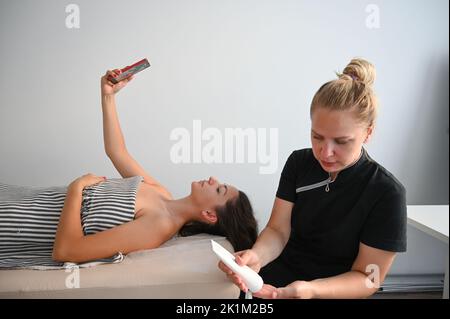 Junge lächelnde Frau beim Selfie im Schönheitssalon. Junge Frau, die Smartphone benutzt, während sie eine Gesichtsmassage im Spa hat. Stockfoto