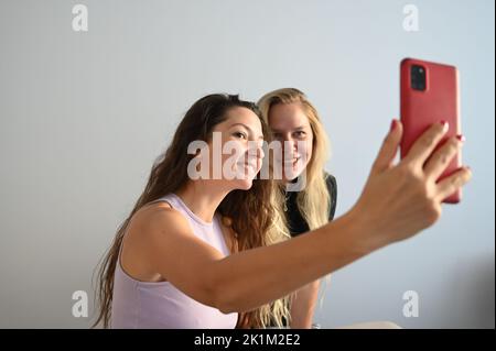 Junge, schmunzelende Frauen machen Selfie im hellen Raum. Porträt von zwei jungen Frauen, die ein Selfie machen Stockfoto
