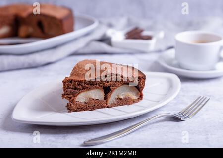 Stück Schokoladenkuchen mit Birne. Im Hintergrund ist eine Torte, eine Tasse Kaffee und Schokoladenstücke. Nahaufnahme. Stockfoto