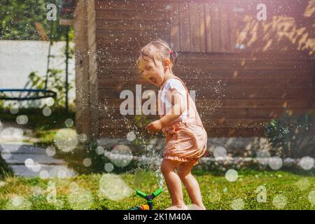 Schönes Mädchen in nasser Kleidung spielen mit Sprinkler im Garten Stockfoto