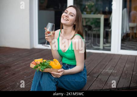 Porträt einer jungen glücklichen, wunderschönen Frau, die auf einer hölzernen Veranda sitzt, ein Glas Weißwein hält und Gemüse schneidet. Stockfoto