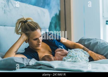 Kaukasische blonde Mutter liegt im Bett mit neugeborenem Baby stillen ihn. Mutterschaft. Säuglingspflege. Verantwortung. Horizontale Innenaufnahme. Hochwertige Fotos Stockfoto