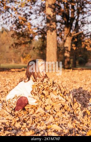 Kleines Mädchen 3-4 Jahre alt hockt im Herbst im Park in den Strahlen der untergehenden Sonne und Abholung große Armvoll von Eichenblättern. Dunkelhaarige Kind in Stockfoto