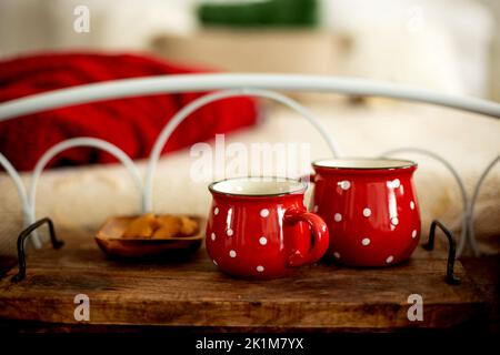 Zwei rote Becher mit weißen Punkten auf einem Tablett mit einem Plätzchen-Teller vor einem Bett in einem weihnachtlich dekorierten Haus Stockfoto