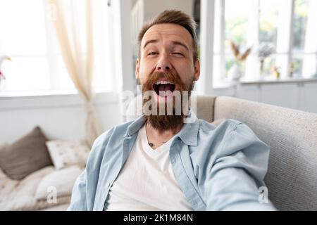Bärtiger, glücklicher junger Mann, der lächelt und Selfie-Fotos macht, während er zu Hause auf dem Sofa sitzt. Stockfoto