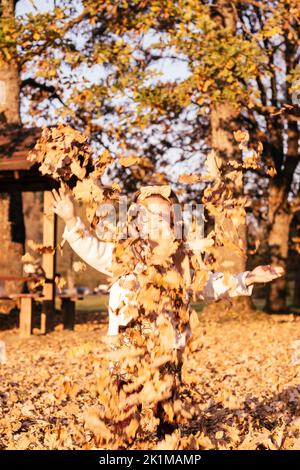 Das kleine Mädchen, 3-4 Jahre alt, steht im Herbstpark in den Strahlen der untergehenden Sonne und wirft Eichenblätter hoch, lächelt und schließt ihre Augen. Dunkelhaarige Kind in Stockfoto