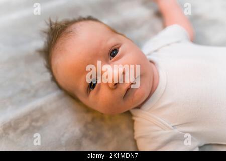 Entzückender dunkelhaariger neugeborener Junge, der mit seinen dunklen Augen intensiv auf die Kamera blickt. Die Neugier des Kindes. Nahaufnahme Innenporträt. Hochwertige Fotos Stockfoto