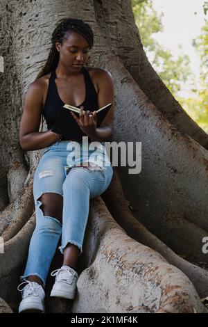 DAS JUNGE AFROAMERIKANISCHE MÄDCHEN RELAXS LIEST, WÄHREND ES IM SCHATTEN EINES BAUMES SITZT. Stockfoto