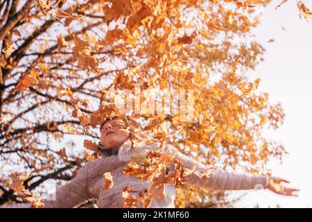 Junge 9-10 Jahre alt steht im Herbstpark in den Strahlen der untergehenden Sonne und wirft Eichenblätter, lächelnd und schließt seine Augen. Defokussiert Kind in grau Stockfoto