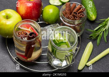 Zimt, Apfel und Gurke infundiert Wasser in Glas. Zimtstangen im Glasgefäß. Gemüse und Obst auf dem Tisch. Schwarzer Hintergrund. Draufsicht Stockfoto