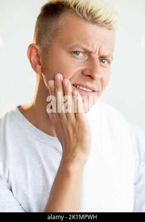 Gesichtspflege für Männer. Nach der Rasur feuchtigkeitsspendende Haut. Morgendliche Routine. Porträt eines gestörten blonden Mannes in weißem T-Shirt, der trockene Wange berührt, isoliert auf einem Seufzer Stockfoto