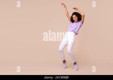 In voller Länge Foto von funky glänzend Mädchen gekleidet lila T-Shirt tanzen leeren Raum isoliert beige Farbe Hintergrund Stockfoto