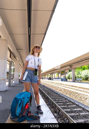 Mädchen wartet auf den Zug am Bahnhof für ihr nächstes Ziel, trägt einen Rucksack, Hut und Sonnenbrille, digitale Nomade und Konzept der Transport und Reise Stockfoto