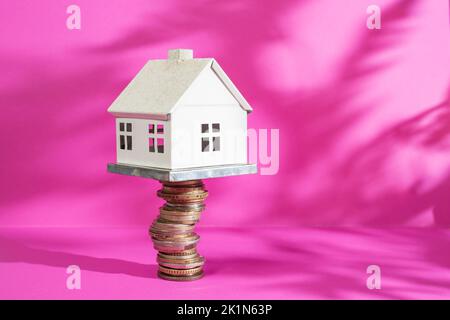 Weißes Modellhaus auf einem Münzstapel, auf magentafarbenem Hintergrund mit Pflanzenschatten. Stockfoto