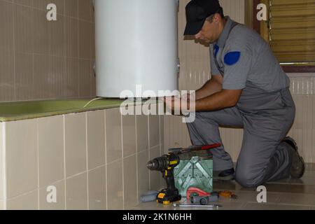 Klempner im Badezimmer eines Hauses, der mit seinen Arbeitswerkzeugen einen elektrischen Warmwasserbereiter repariert. Installation eines Boilers im Badezimmer, Do-it-yourself Stockfoto