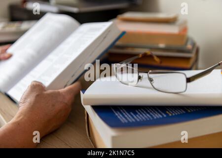 Eine Brille auf einem Schreibtisch mit einer Person, die ein Buch liest. Stockfoto