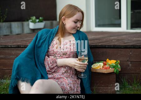 Nachdenkliche, romantische, ruhige blonde Frau, Studentin in pinkem Kleid, blaue Decke mit Weinglas und Obstteller auf der Terrasse bedecken Stockfoto