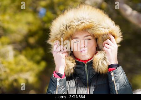 Junge Frau in einer pelzigen Kapuzenjacke, die einen Kuss in einen Wald bläst. Stockfoto