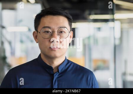 Nahaufnahme Porträt des erfolgreichen asiatischen Investors in Brille und blauem Hemd, Geschäftsmann, der nachdenklich auf die Kamera schaut, Mann, der im modernen Bürogebäude arbeitet, erfahrener und reifer Chef-Manager. Stockfoto