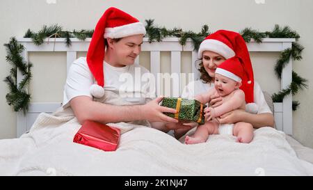 Frau und Mann schenken dem Baby auf dem Bett, das zu weihnachten und Neujahr dekoriert ist, ein Familienpaar im Schlafzimmer zu Hause Stockfoto