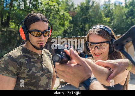 Langhaarige schöne Mädchen mit Kopfhörern und Schutzbrille üben Ziel mit Instruktoren auf dem Schießstand, mittlere Nahaufnahme. Hochwertige Fotos Stockfoto