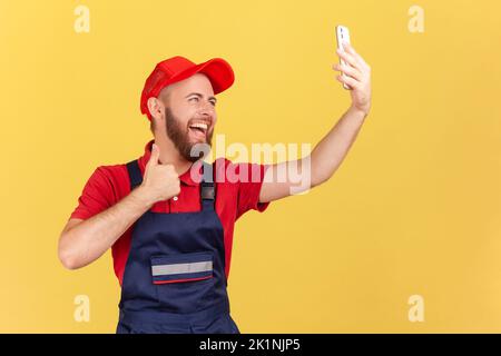 Porträt eines positiven Mitarbeiters in Overalls, der Selfie macht oder bei einem Videoanruf spricht, auf die Gerätekamera schaut und dem Kunden den Daumen zeigt. Innenaufnahme des Studios isoliert auf gelbem Hintergrund. Stockfoto