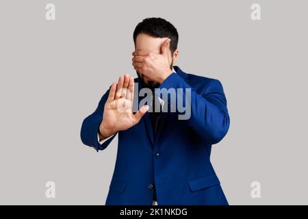 Mann, der die Augen mit der Hand schließt, die Handfläche ausstreckt, will das nicht sehen, Probleme ignorieren, sich vor stressigen Situationen verstecken, einen offiziellen Anzug tragen. Innenaufnahme des Studios isoliert auf grauem Hintergrund. Stockfoto