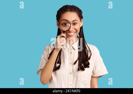 Porträt einer lustigen Frau mit schwarzen Dreadlocks, die Lupe hält und mit großem Zoomauge, positivem Gesicht und weißem Hemd auf die Kamera schaut. Innenaufnahme des Studios isoliert auf blauem Hintergrund. Stockfoto