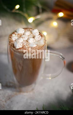 Heiße Schokolade in einem Glas-Becher mit Mini-Marschmalchen auf grauem Tisch, unscharfes Licht im Hintergrund Stockfoto