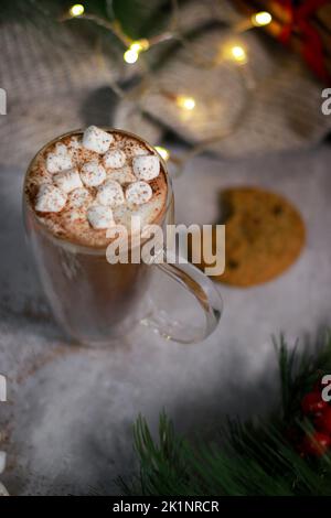 Draufsicht auf heiße Schokolade in einem Glaskrug mit Mini-Marschmalven auf grauem Tisch, verschwommener Weihnachtshintergrund Stockfoto