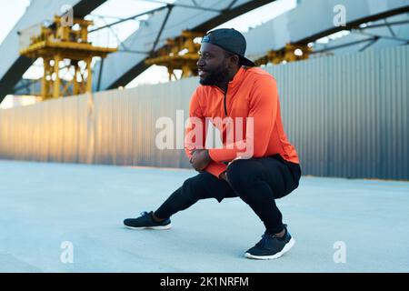 Glücklicher junger afroamerikanischer Sportler im aktiven Ohr, der Stretching-Übungen mit seinem linken Bein im Knie im städtischen Umfeld macht Stockfoto