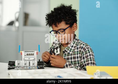 Clever kleiner Junge in Casualwear und Brillen verbindet Details des Spielzeugroboters, während er am Schreibtisch sitzt und individuell arbeitet Stockfoto