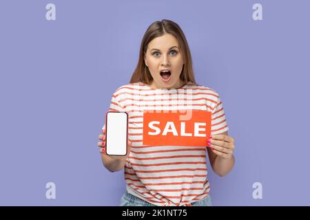 Porträt einer erstaunt überraschten blonden Frau in einem gestreiften T-Shirt mit Verkaufskarte und Smartphone mit leerem Display, Platz zum Werben. Innenaufnahme des Studios isoliert auf violettem Hintergrund. Stockfoto