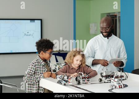 Glücklicher junger Roboterlehrer mit Tablet, der neben zwei cleveren interkulturellen Schülern steht, die mit neuen Robotermodellen spielen Stockfoto