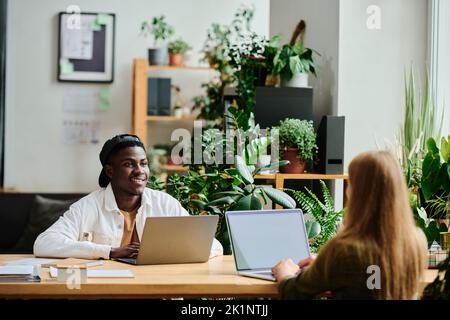 Junger glücklicher schwarzer Mann in Casualwear, der eine blonde Kollegin mit einem Laptop vor ihm sitzend ansieht und die Präsentation vorbereitet Stockfoto
