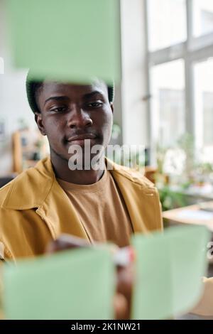 Junger, seriöser Büroleiter, der vor einer transparenten Tafel steht und Notizen auf Notizbüchern macht, während er seine Arbeit plant Stockfoto