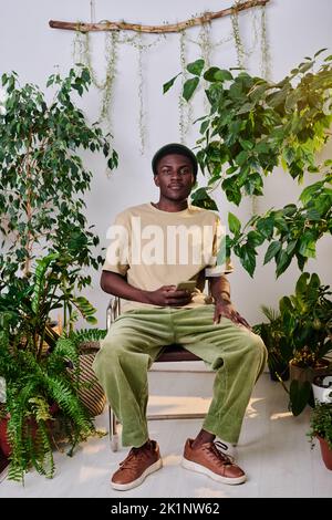 Junger afroamerikanischer Mann in Casualwear mit Smartphone, das auf einem Stuhl zwischen grünen Pflanzen im Büro sitzt und die Kamera anschaut Stockfoto