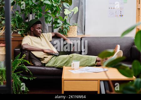 Junger entspannter Mann in Casualwear mit Laptop auf den Knien, der auf der schwarzen Ledercouch sitzt und online Daten für ein neues Projekt durchschaut Stockfoto