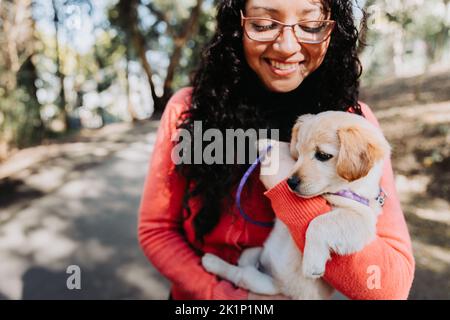 Lateinische lockige Brünette Frau, umarmt und hält mit einem goldenen Retriever Welpen im Park. Stockfoto