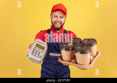Freundlicher, optimistischer Lieferer, der Terminal und Kaffee mit Pizzakarton hält, die Kamera mit fröhlichem Ausdruck anschaut, Overalls und Mütze trägt. Innenaufnahme des Studios isoliert auf gelbem Hintergrund. Stockfoto