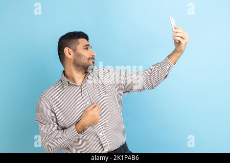 Fröhlich lächelnder Geschäftsmann Blogger, der auf die Smartphone-Kamera schaut, mit den Abonnenten kommuniziert, streamt, gestreiftes Hemd trägt. Innenaufnahme des Studios isoliert auf blauem Hintergrund. Stockfoto