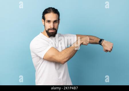 Porträt eines ungeduldigen, wütenden Mannes mit Bart und weißem T-Shirt, der auf die Armbanduhr zeigt und Unzufriedenheit mit verspäteter Besprechung und verzögerter Zeit zum Ausdruck bringt. Innenaufnahme des Studios isoliert auf blauem Hintergrund. Stockfoto